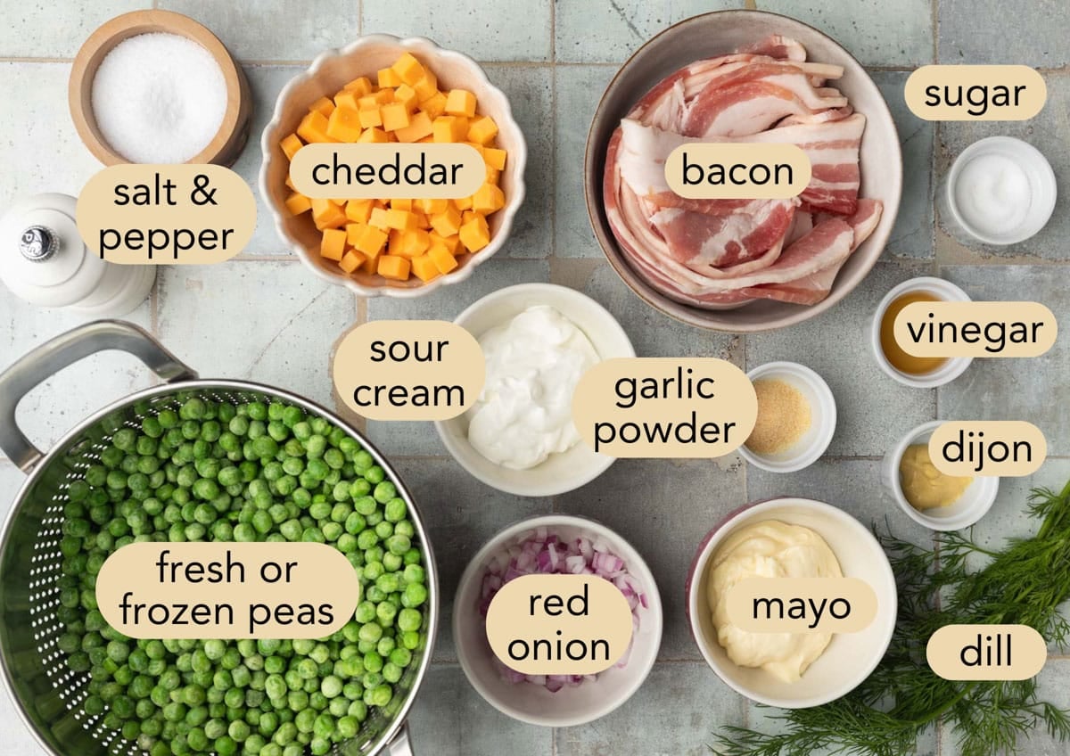 Ingredient shot for creamy pea salad arranged on a blue-gray tiled surface. The labeled ingredients include fresh or frozen peas, bacon, cheddar cheese, mayo, sour cream, red onion, dill, Dijon mustard, vinegar, garlic powder, sugar, and salt and pepper