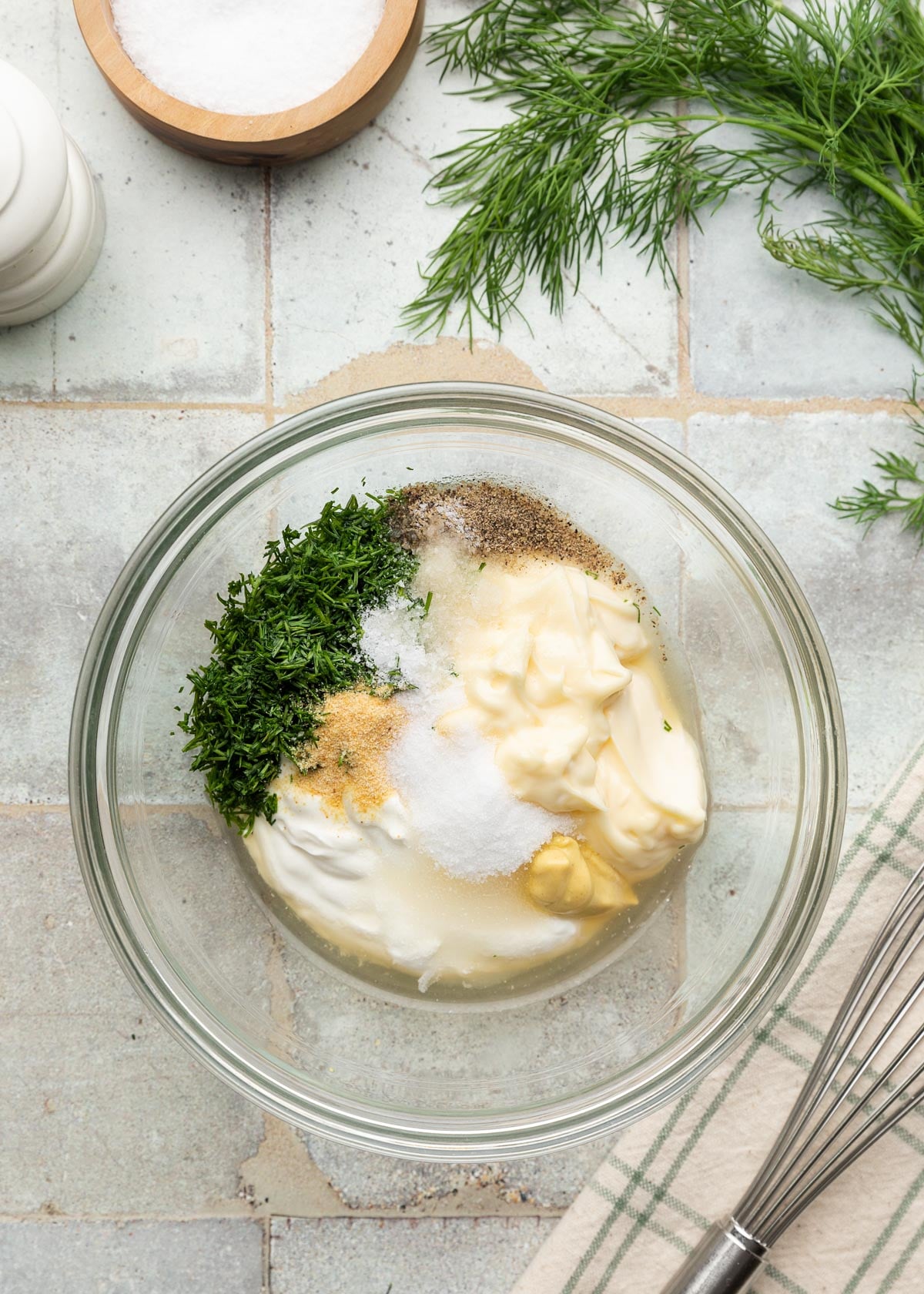 Top-down photo of ingredients for a creamy dill dressing in a glass mixing bowl, including mayonnaise, sour cream, Dijon mustard, vinegar, chopped fresh dill, sugar, garlic powder, salt, and black pepper. A whisk, salt cellar, and fresh dill are arranged around the bowl on a tiled surface.