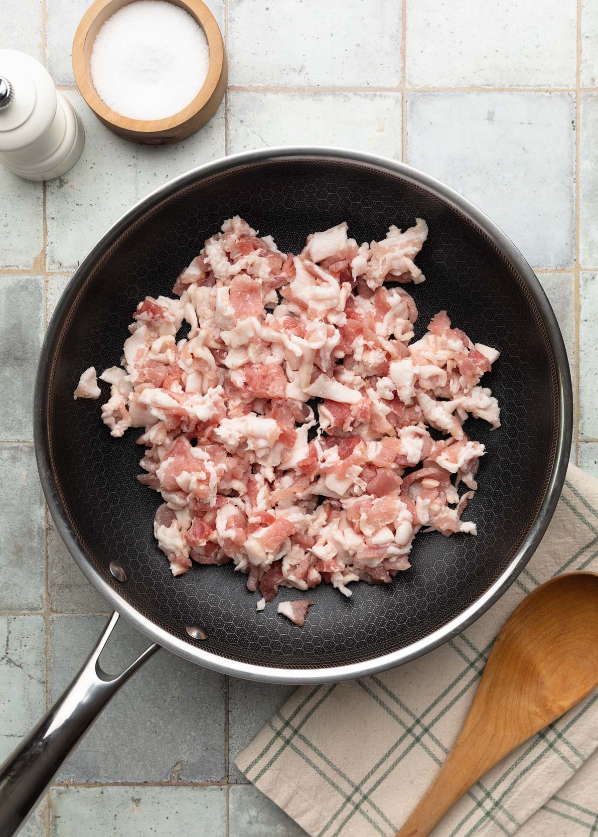Top-down photo of raw chopped bacon spread in a skillet before cooking, with a wooden spoon and seasonings nearby on a tiled surface.