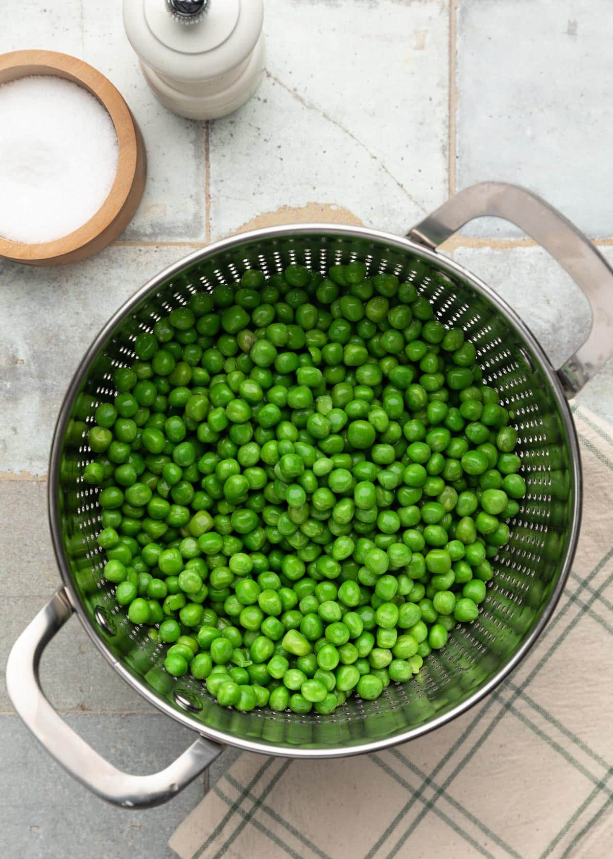 Top-down photo of bright green peas in a metal colander after blanching, set on a tiled surface with salt and pepper nearby.