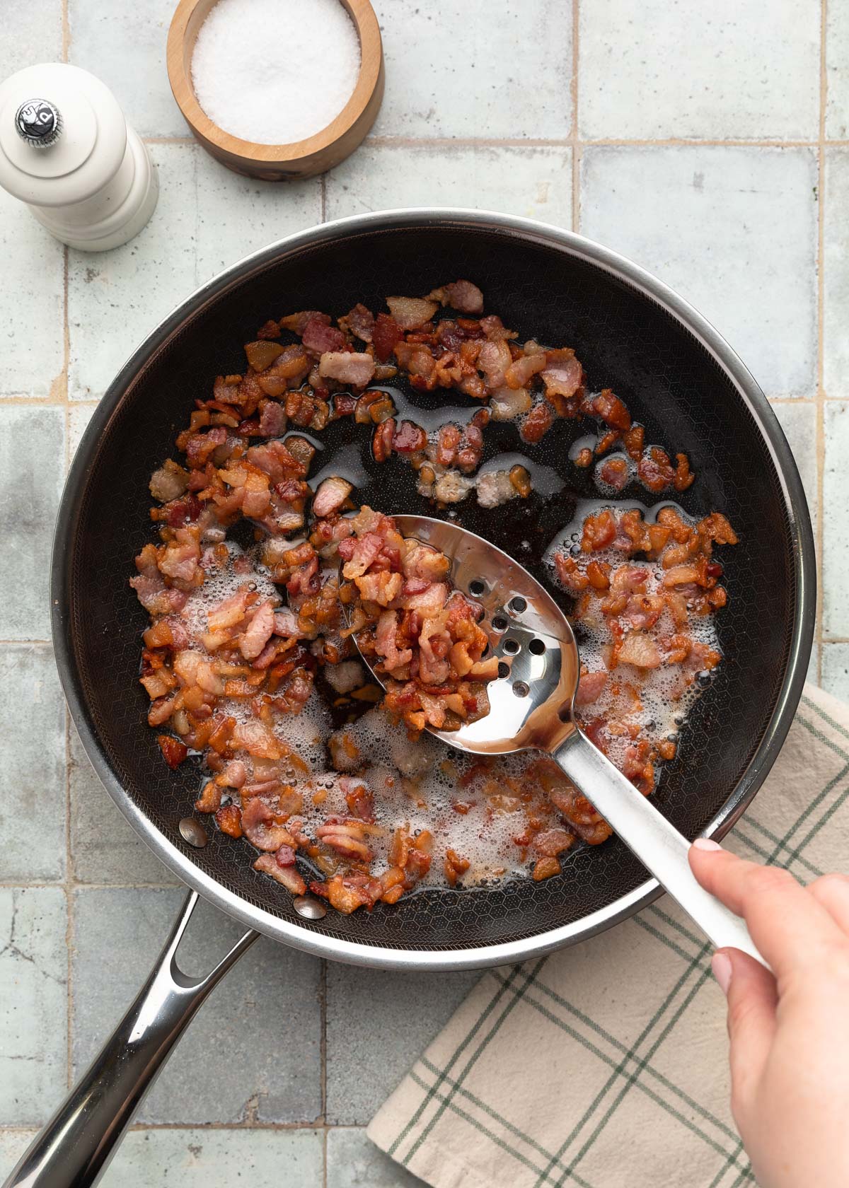 Top-down process photo of chopped bacon cooking in a skillet, with a slotted spoon lifting up crispy browned pieces from the rendered bacon fat.