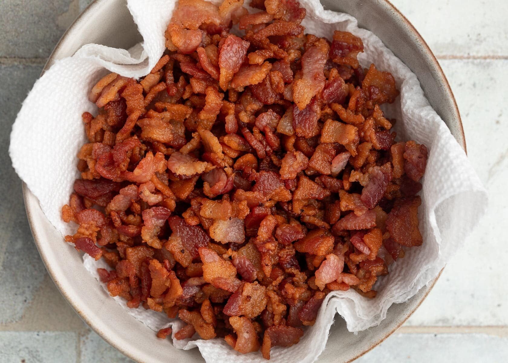 Top-down photo of crispy chopped bacon draining on a paper towel-lined bowl after cooking.