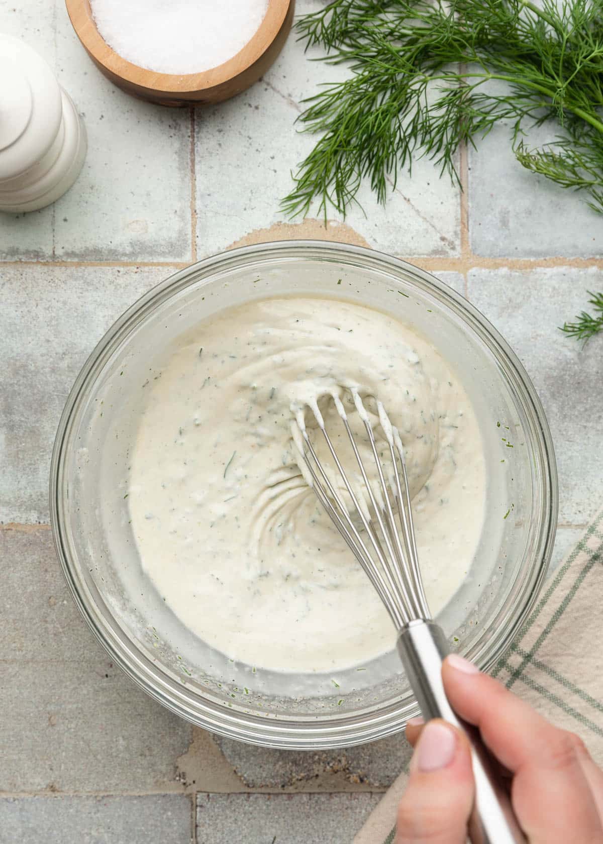 Top-down process photo of a creamy dill dressing being whisked together in a glass mixing bowl, with fresh dill and seasonings on the tiled surface nearby.