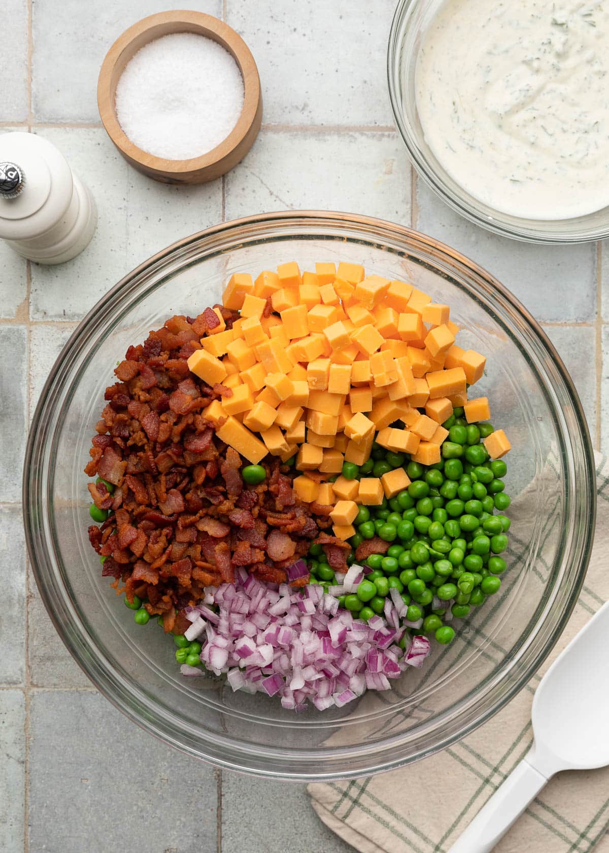 Top-down ingredient photo showing green peas, crispy chopped bacon, cheddar cheese cubes, and diced red onion arranged in a glass mixing bowl before tossing the salad together, with a separate bowl of creamy dill dressing beside it on the tiled surface.