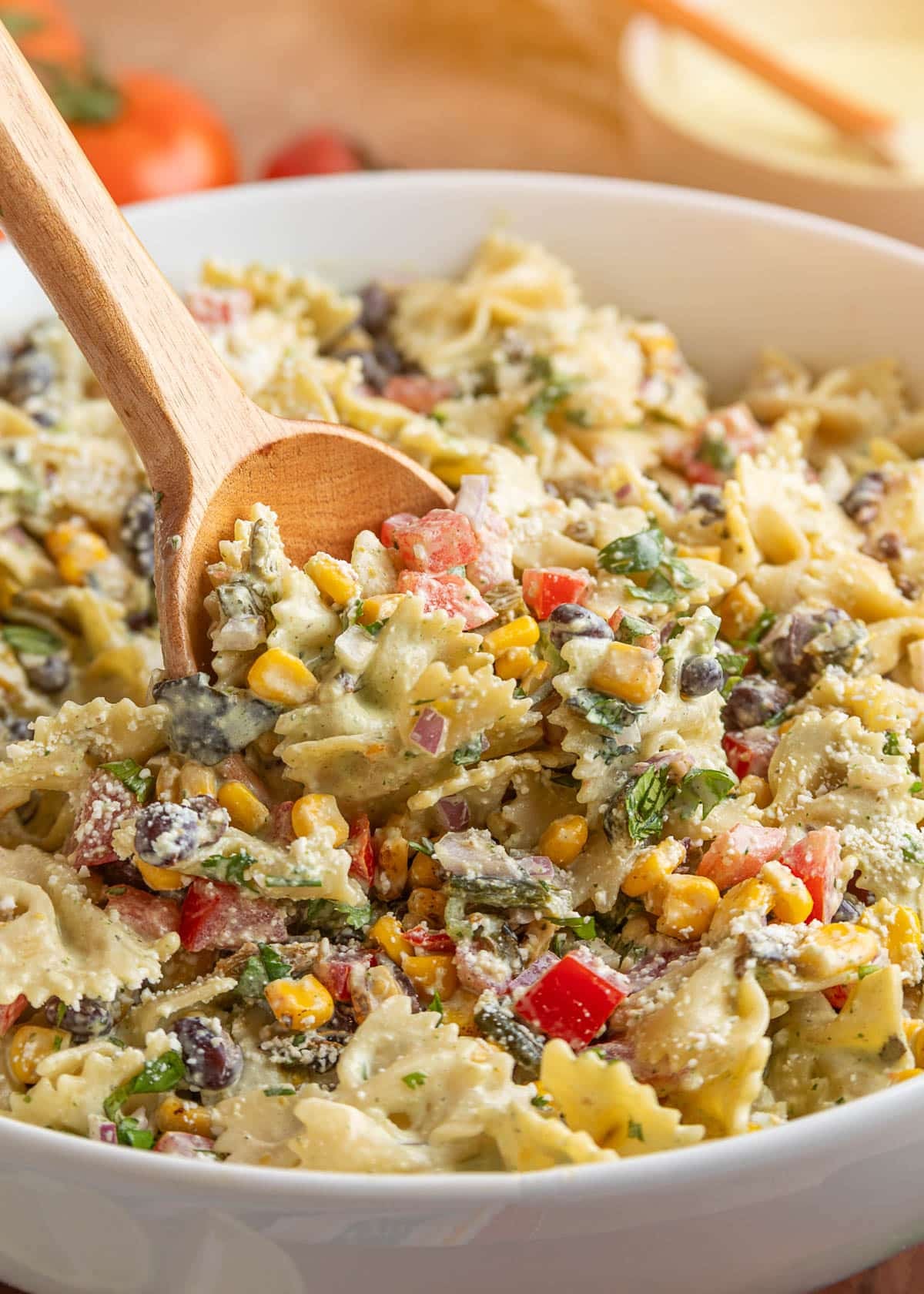 Wooden spoon stirring creamy Southwest pasta salad with farfalle pasta, roasted corn, black beans, tomatoes, poblano peppers, cotija cheese, and cilantro jalapeno dressing.