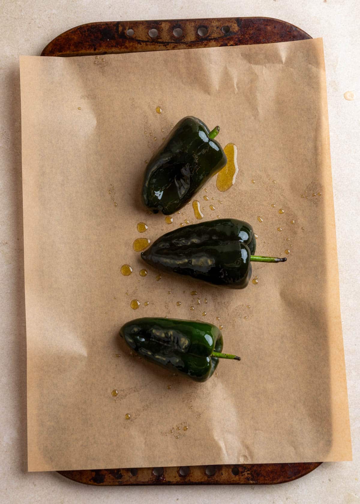 Overhead photo of whole poblano peppers drizzled with oil on a parchment-lined baking sheet, ready for roasting.