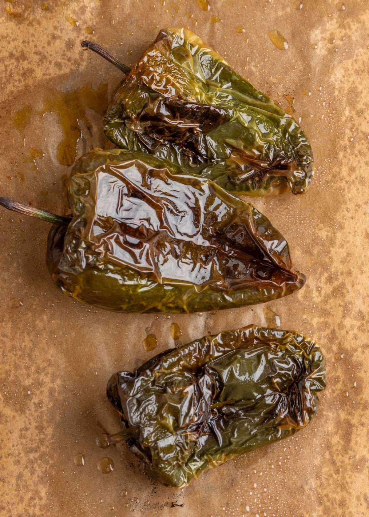 Close-up overhead photo of roasted poblano peppers with blistered, charred skins on a parchment-lined baking sheet after roasting.