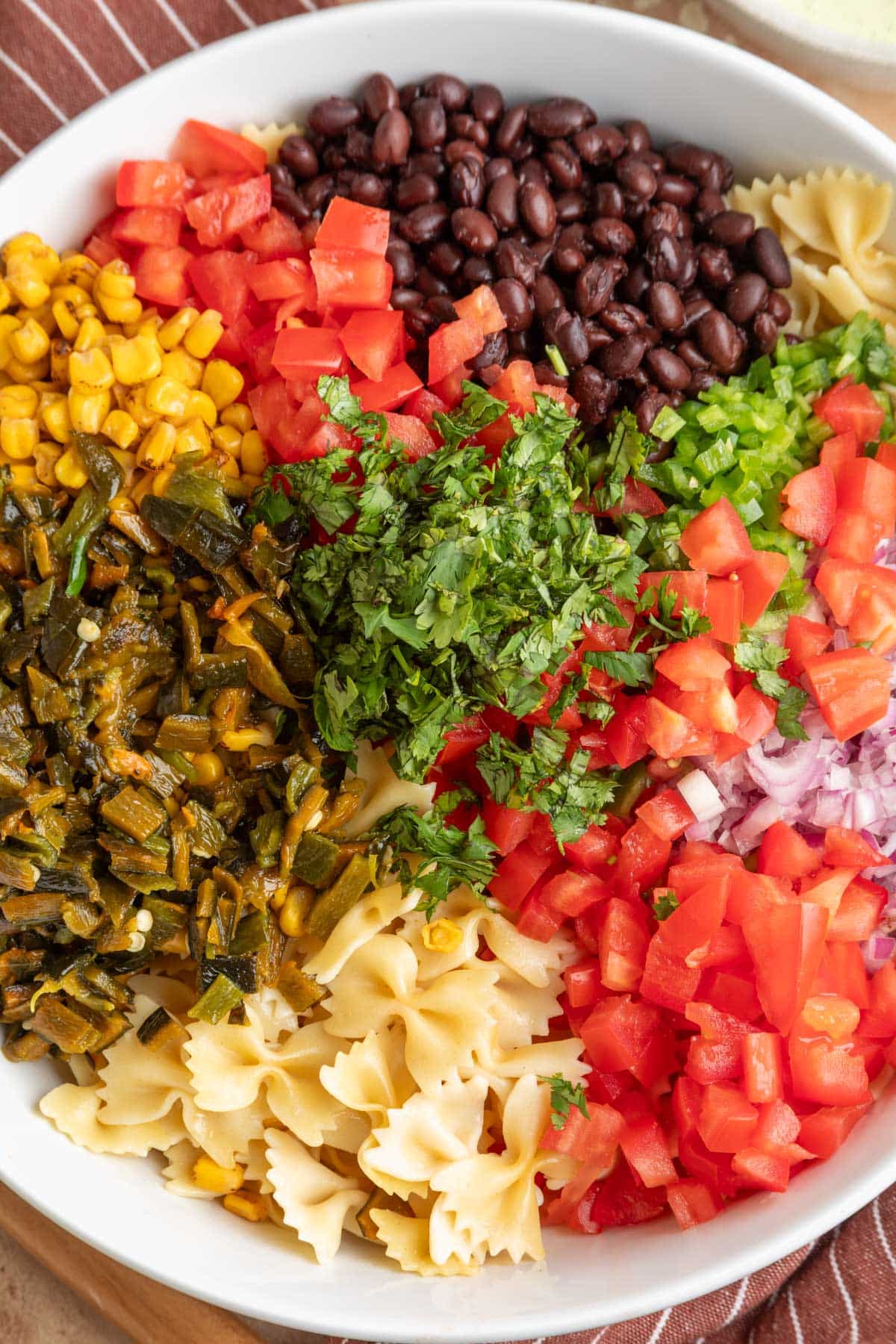 Overhead photo of Southwest pasta salad ingredients layered in a white serving bowl with farfalle pasta, black beans, diced Roma tomatoes, roasted poblano peppers, fire roasted corn, red onion, jalapeno, and fresh cilantro before tossing.