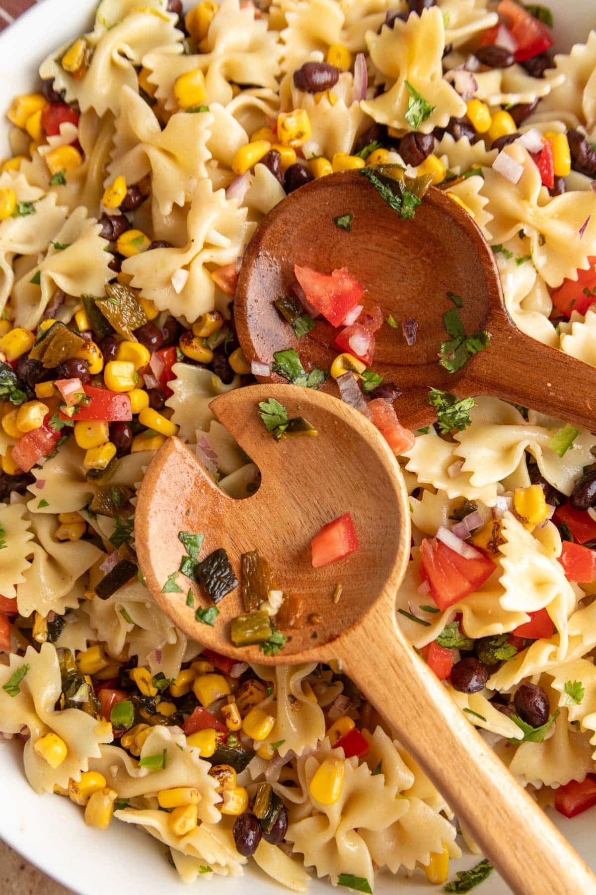 Close-up of Southwest pasta salad being tossed with wooden salad servers, featuring farfalle pasta, black beans, roasted corn, tomatoes, roasted poblano peppers, red onion, and cilantro.