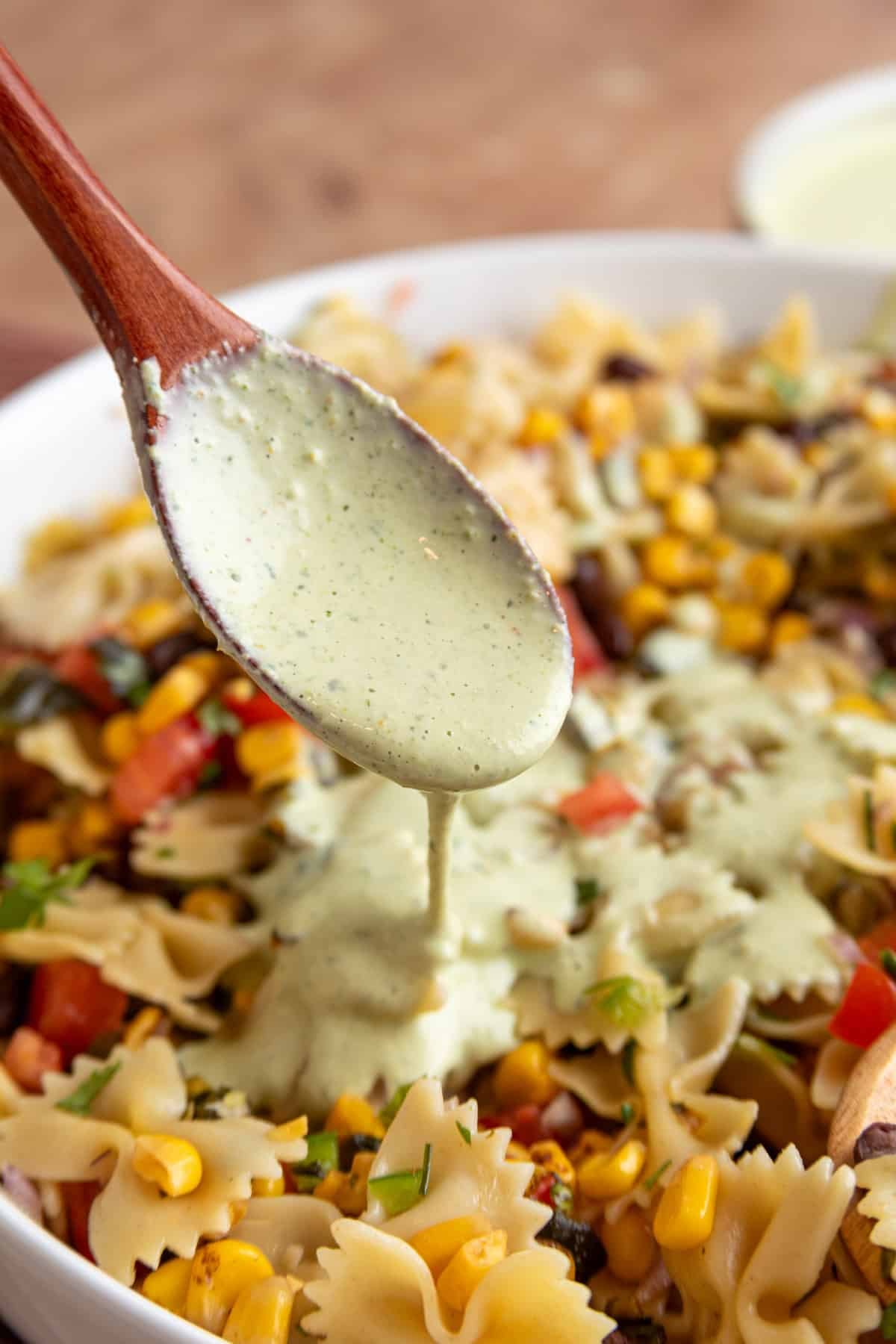 Wooden spoon drizzling creamy cilantro jalapeno dressing over Southwest pasta salad with farfalle pasta, black beans, roasted corn, diced tomatoes, and fresh herbs.