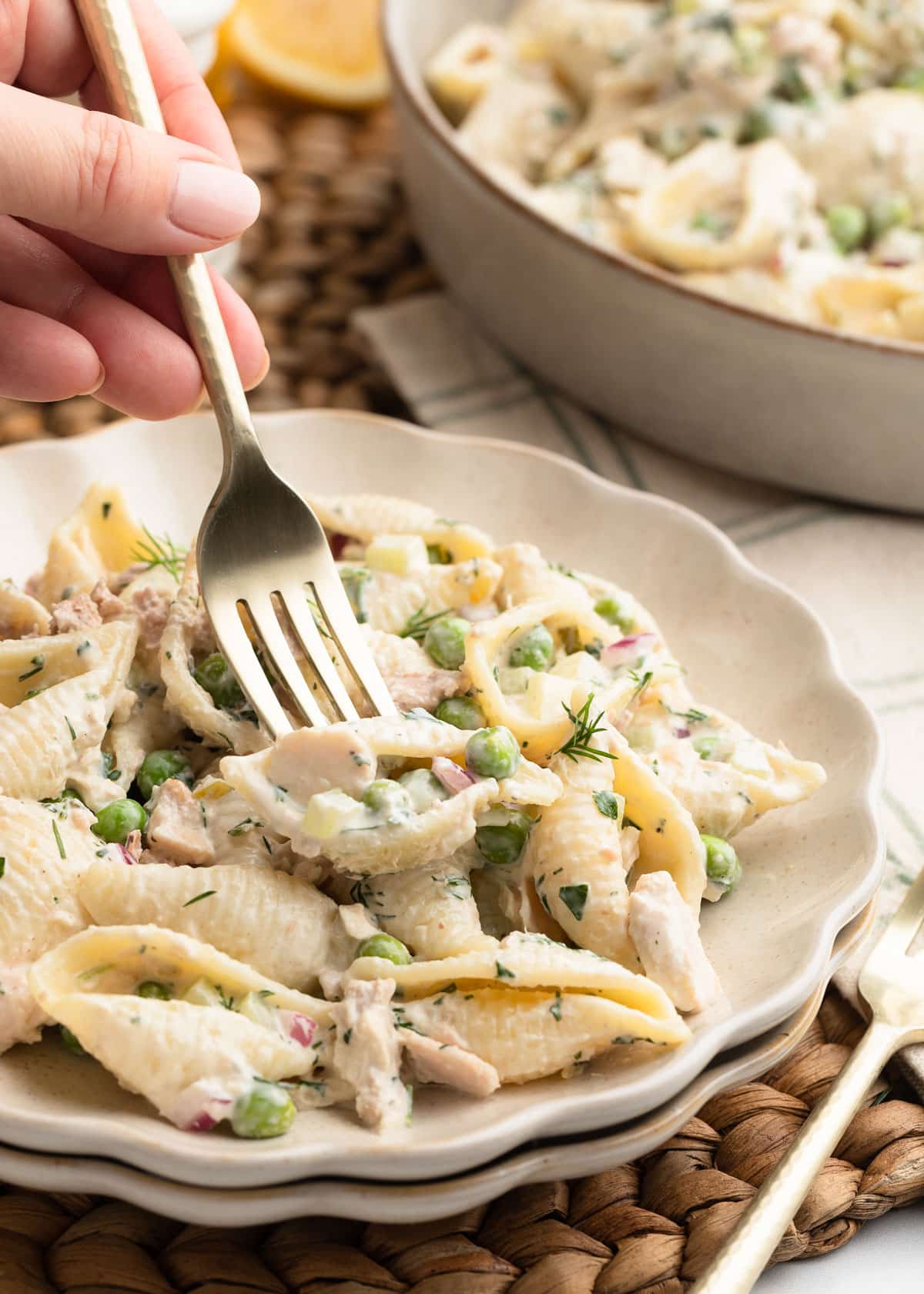 Close-up photo of a plated serving of creamy tuna pasta salad with shell pasta, tuna, peas, celery, red onion, and dill in a creamy dressing. A hand holds a fork over the bowl, with a larger serving dish blurred in the background on a woven placemat.