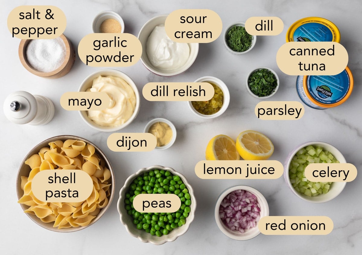 Ingredients for tuna pasta salad including shell pasta, canned tuna, peas, celery, red onion, mayo, sour cream, dill relish, Dijon mustard, lemon juice, fresh dill, parsley, and seasonings.