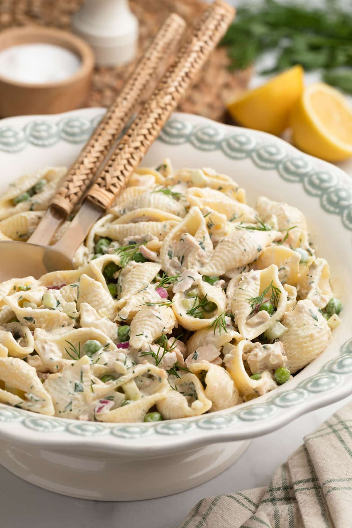 Styled serving photo of creamy tuna pasta salad in a decorative white serving bowl with woven-handled serving utensils resting on top. The salad contains shell pasta, tuna, peas, celery, red onion, and fresh dill in a creamy herb dressing, with lemons and herbs softly blurred in the background.
