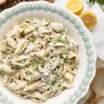 Overhead serving photo of creamy tuna pasta salad in a decorative white serving bowl with a pale green patterned rim. The salad is made with shell pasta, tuna, green peas, celery, red onion, and fresh dill in a creamy herb dressing, with lemon halves, fresh herbs, and wooden salad servers styled around the bowl.