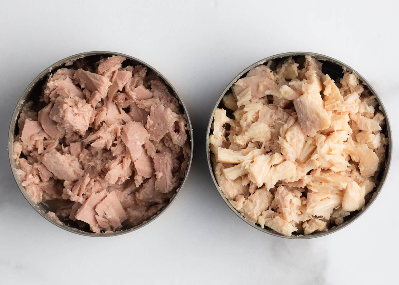 Overhead close-up of two opened cans of tuna on a white marble background. One can contains lighter-colored albacore tuna, while the other contains darker skipjack tuna packed in larger flakes and pieces.
