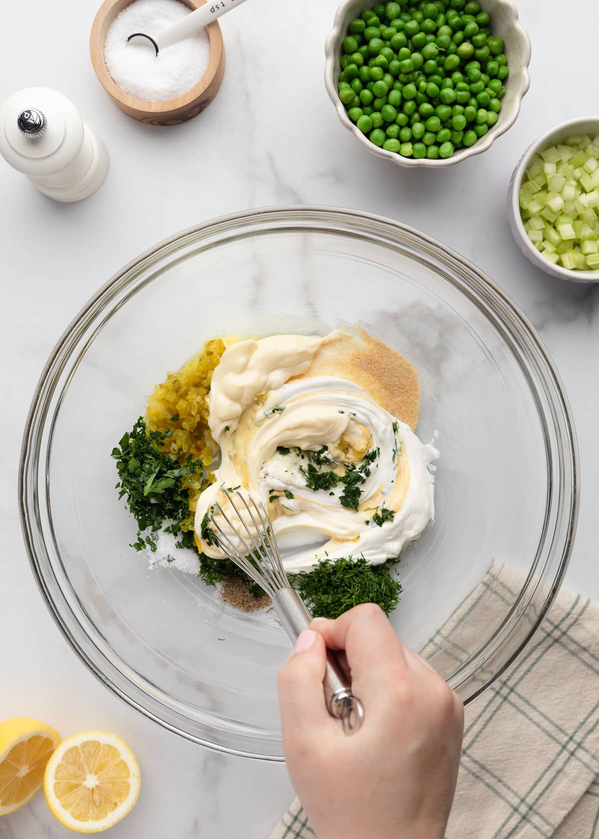Overhead step-by-step photo of tuna pasta salad dressing ingredients in a large glass bowl before mixing. Mayo, sour cream, chopped dill, parsley, dill relish, garlic powder, salt, and pepper are being whisked together, with bowls of peas and celery nearby on a white marble background.