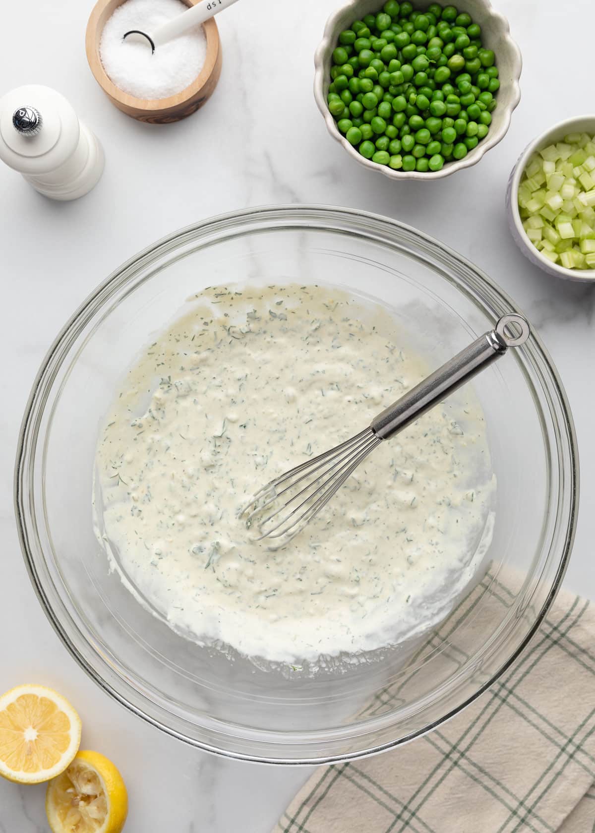 Overhead photo of a creamy tuna pasta salad dressing whisked together in a large glass mixing bowl on a white marble surface. The dressing is speckled with fresh dill and surrounded by bowls of green peas, diced celery, lemons, salt, and pepper.