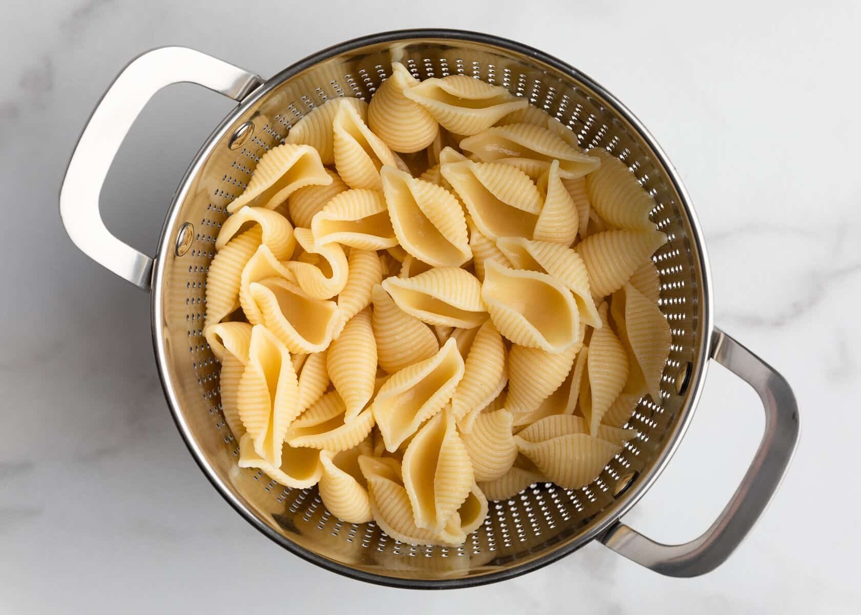 Overhead photo of cooked shell pasta draining in a stainless steel colander on a white marble surface. The pasta shells are pale golden and glossy from cooking, with the ridged texture clearly visible.