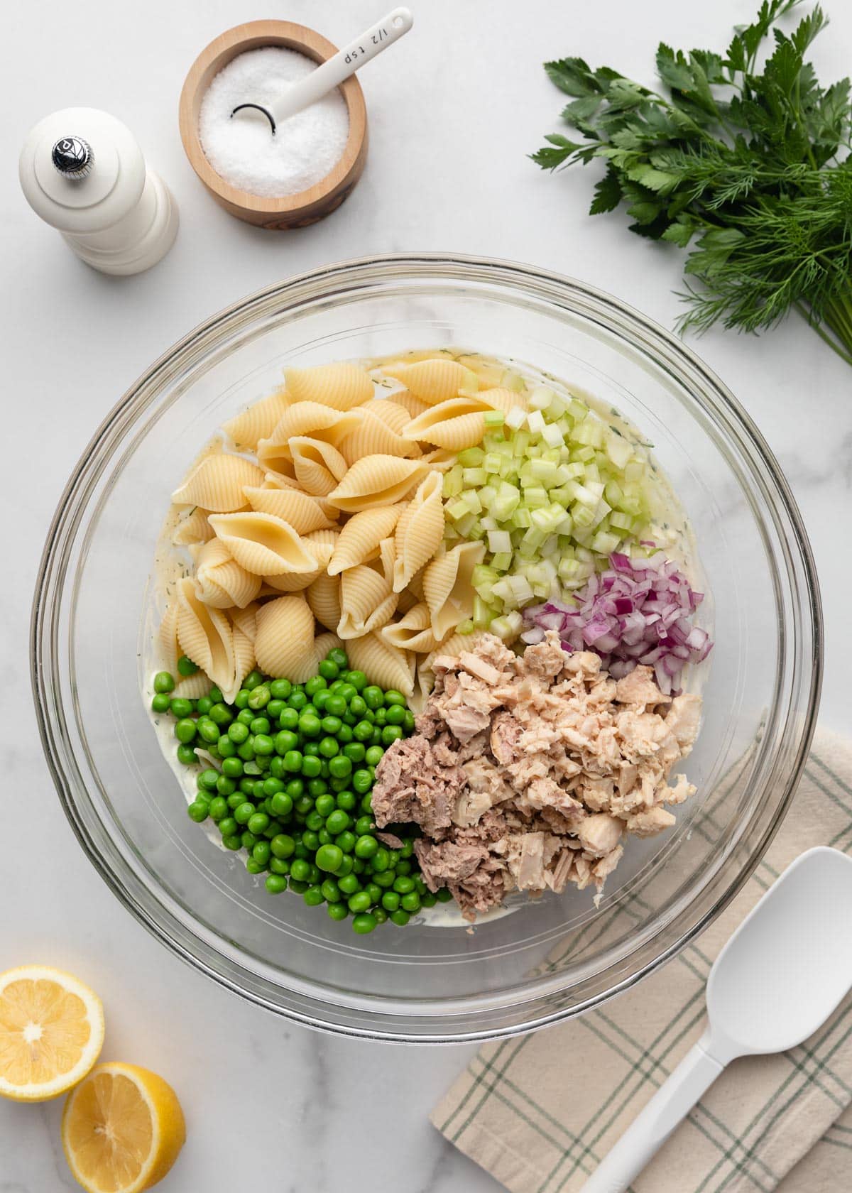 Overhead step-by-step photo of tuna pasta salad ingredients added to a large glass mixing bowl. Cooked shell pasta, green peas, canned tuna, diced celery, and red onion sit on top of a creamy dill dressing, with fresh herbs, lemons, salt, and pepper arranged around the bowl on a white marble surface.