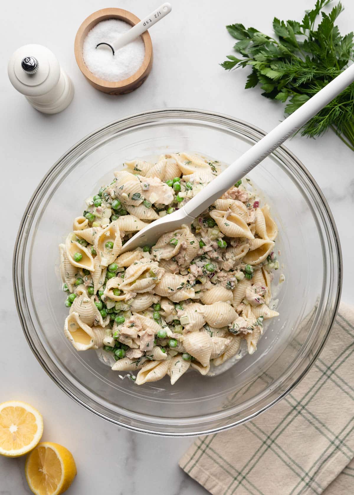 Overhead photo of creamy tuna pasta salad being mixed in a large glass bowl with a white spatula. The salad contains shell pasta, tuna, green peas, diced celery, red onion, and fresh dill coated in a creamy herb dressing, with lemons, parsley, dill, salt, and pepper nearby on a white marble background.