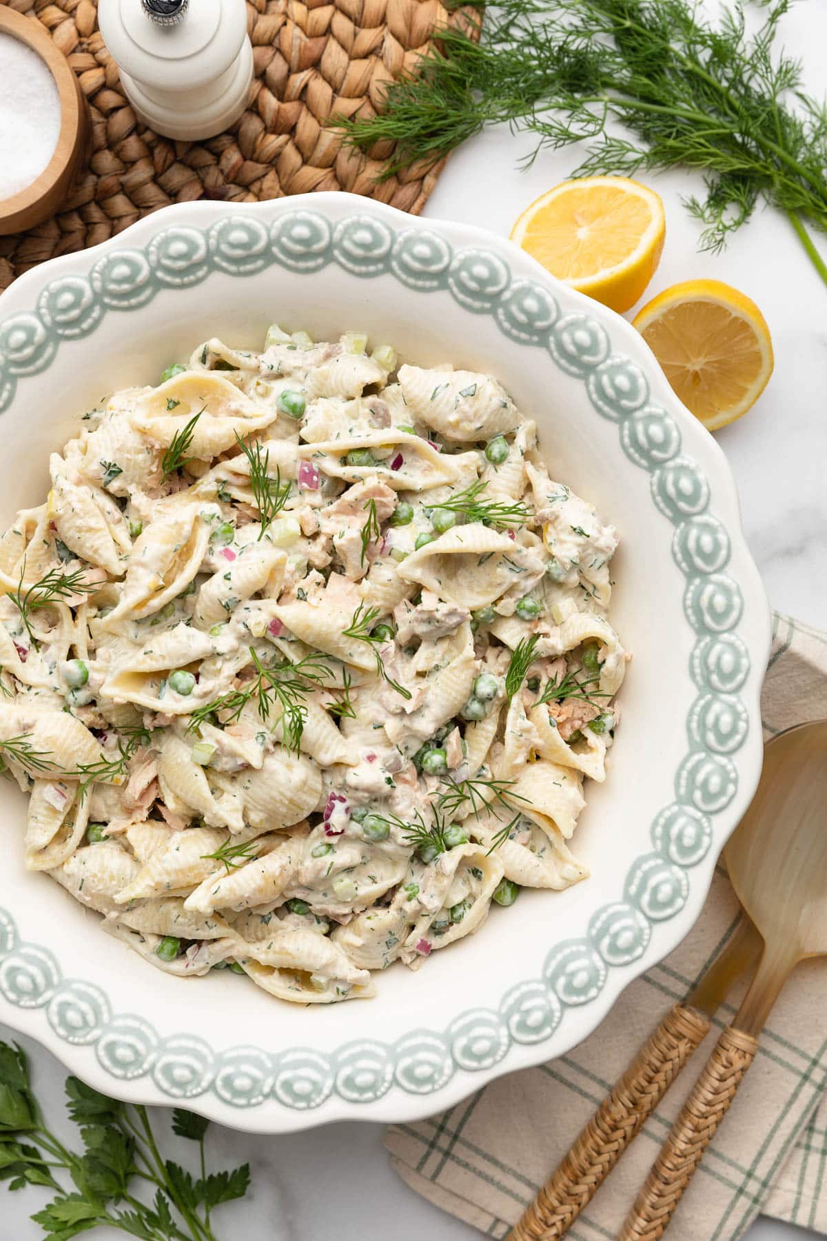Overhead photo of a large white serving bowl filled with creamy tuna pasta salad made with shell pasta, tuna, peas, celery, red onion, and fresh dill. The bowl sits on a white marble surface surrounded by lemon halves, fresh dill, a salt cellar, pepper grinder, and wooden salad servers on a striped cloth napkin.