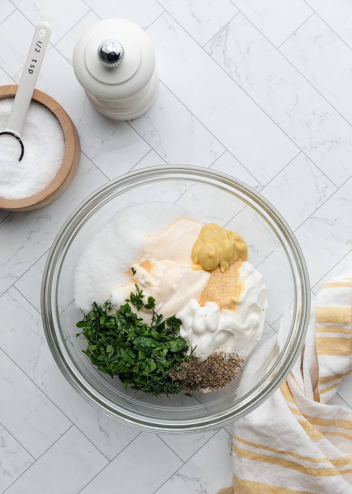 Overhead view of a glass mixing bowl filled with pasta salad dressing ingredients, including mayonnaise, sour cream, Dijon mustard, sugar, garlic powder, chopped fresh herbs, salt, and black pepper, arranged on a white marble-patterned surface beside a pepper mill and striped kitchen towel.