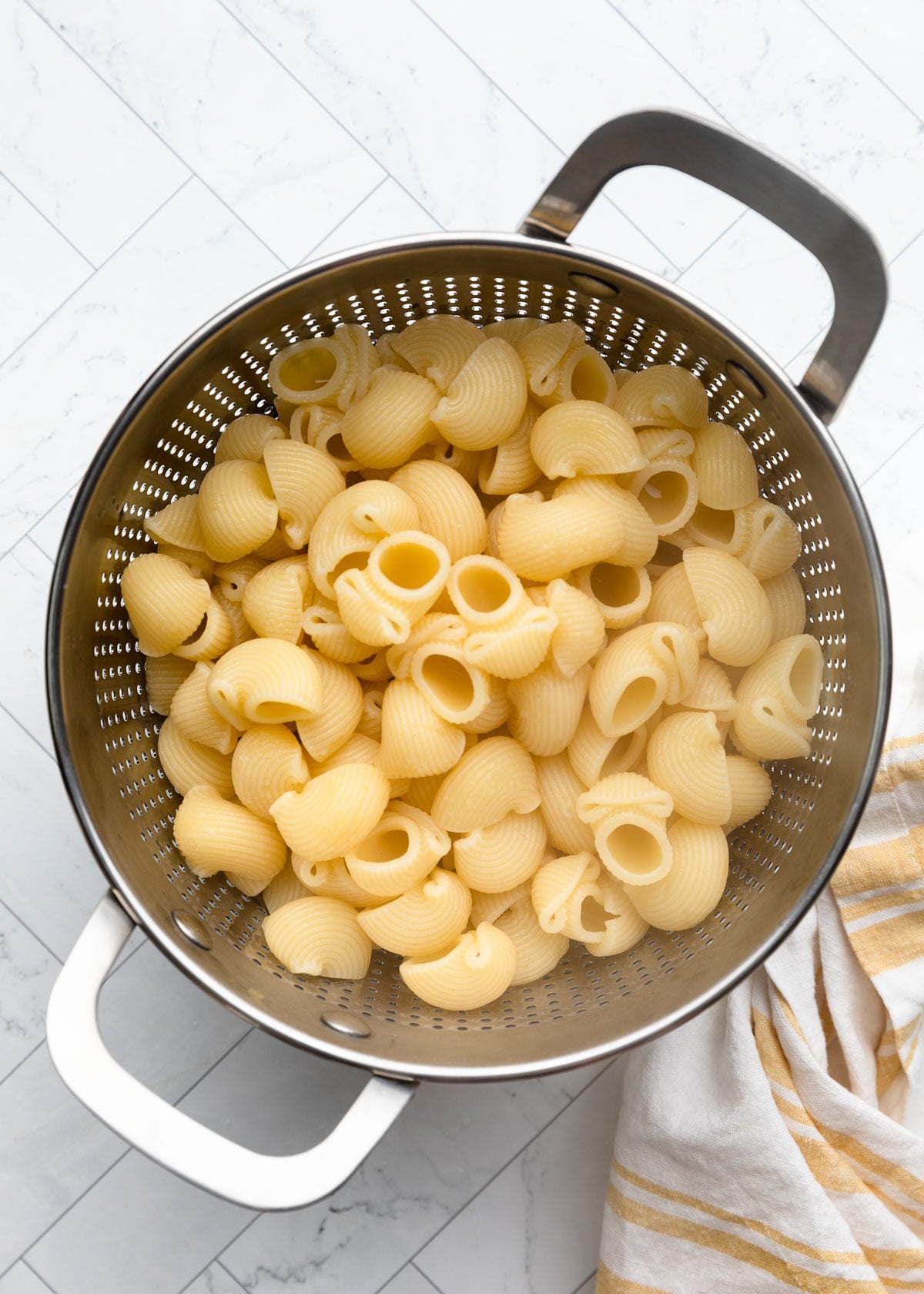 Close-up overhead view of freshly cooked pipette pasta draining in a stainless steel colander, with a striped kitchen towel beside it on a white marble-patterned surface.