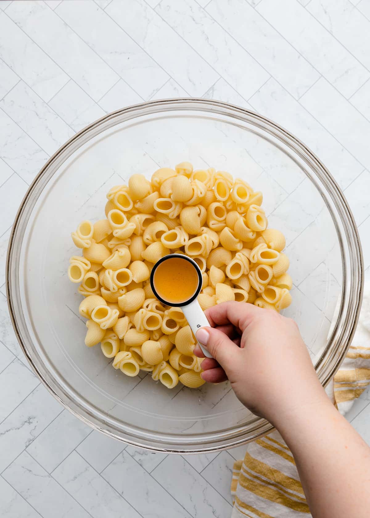 Overhead view of cooked pipette pasta in a large glass mixing bowl while a hand pours vinegar from a measuring spoon over the pasta. A striped kitchen towel rests beside the bowl on a white marble-patterned surface.