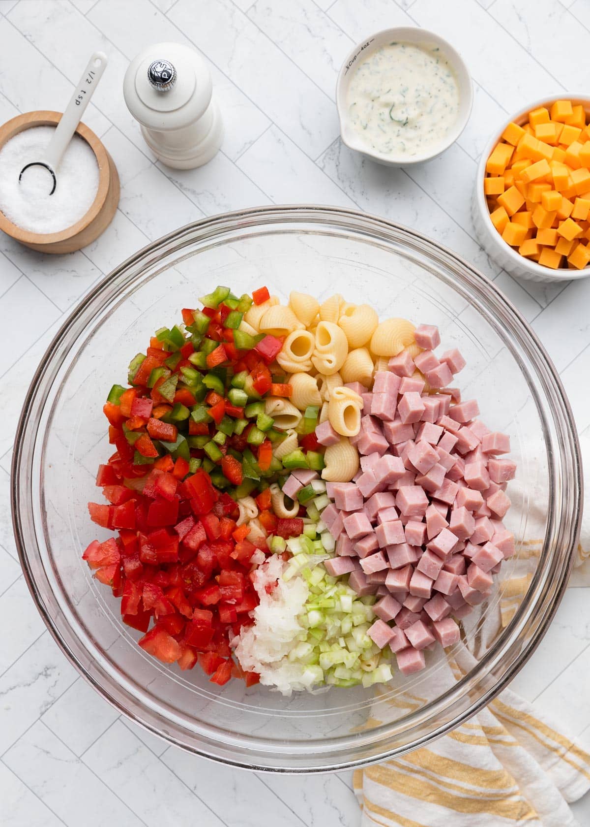 Overhead view of a large glass bowl filled with cooked pipette pasta, diced ham, chopped red and green bell peppers, diced tomato, grated onion, and celery, with a bowl of cheddar cheese cubes and a pitcher of creamy herb dressing nearby.