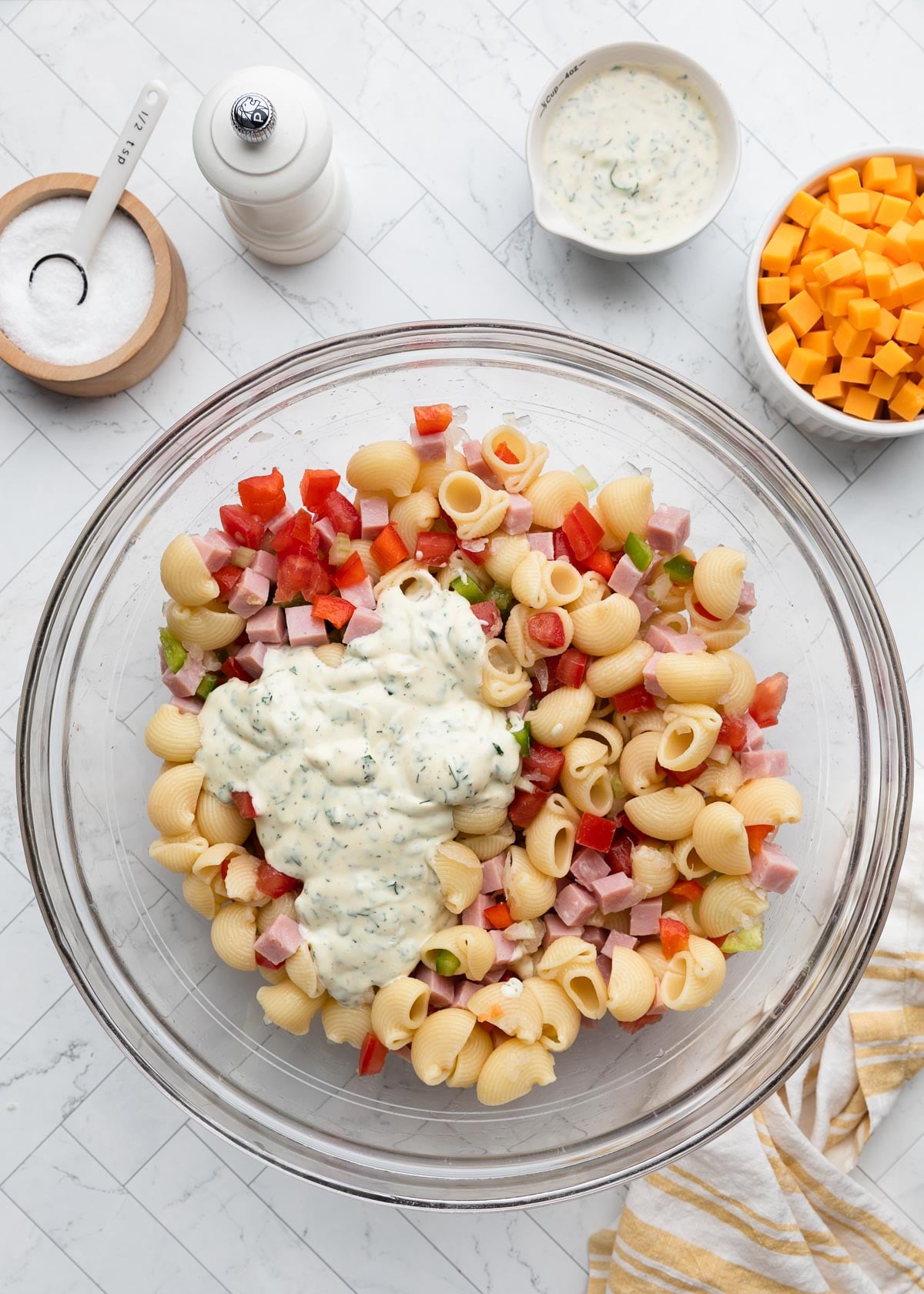 Overhead view of ham pasta salad ingredients tossed together in a glass bowl, topped with creamy herb dressing before mixing, with cheddar cheese cubes and extra dressing visible alongside the bowl.