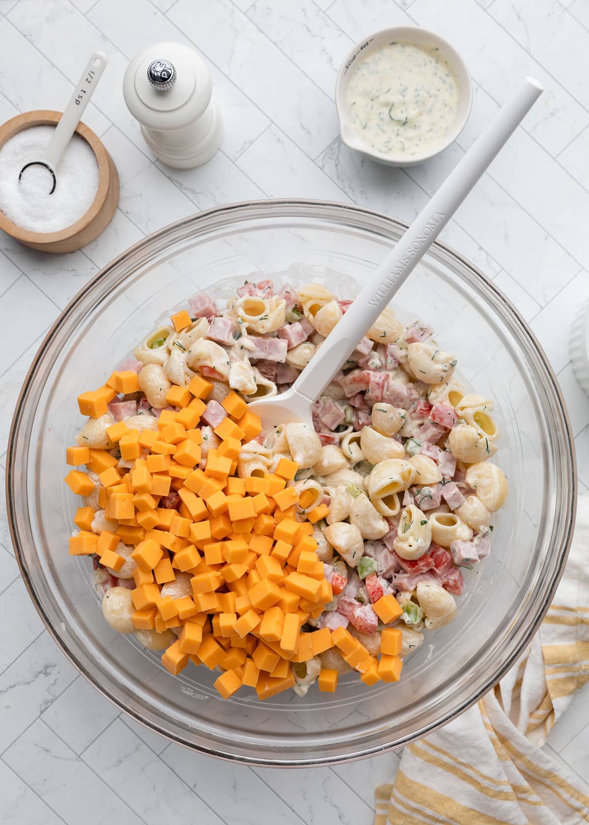 Overhead view of a glass bowl filled with creamy ham pasta salad ingredients, with cubes of cheddar cheese just added on top of dressed pasta, diced ham, tomatoes, peppers, and celery. A pitcher of herb dressing, salt cellar, and pepper mill sit nearby.