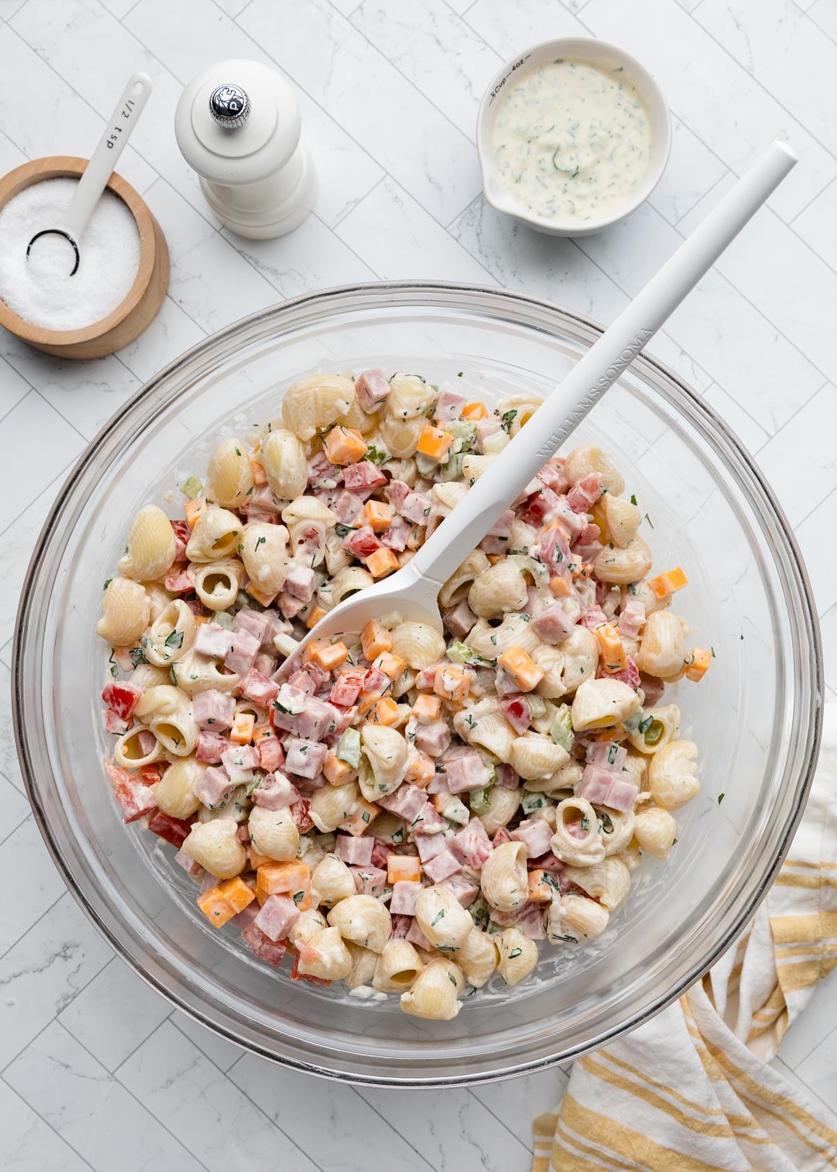 Overhead view of assembled ham pasta salad tossed with cheddar cheese cubes, diced ham, pasta shells, tomatoes, peppers, celery, and creamy herb dressing in a large glass mixing bowl with a rubber spatula.