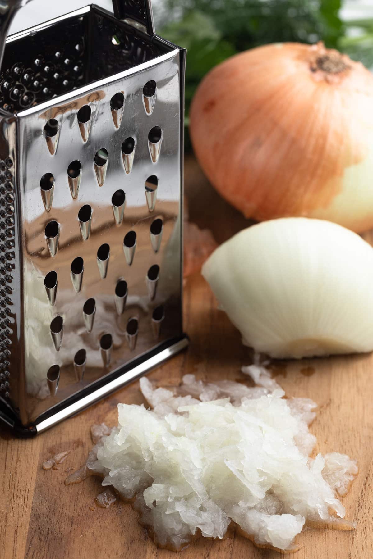 Close-up of a box grater and grated onion on a wooden cutting board, with a halved sweet onion and fresh herbs in the background.