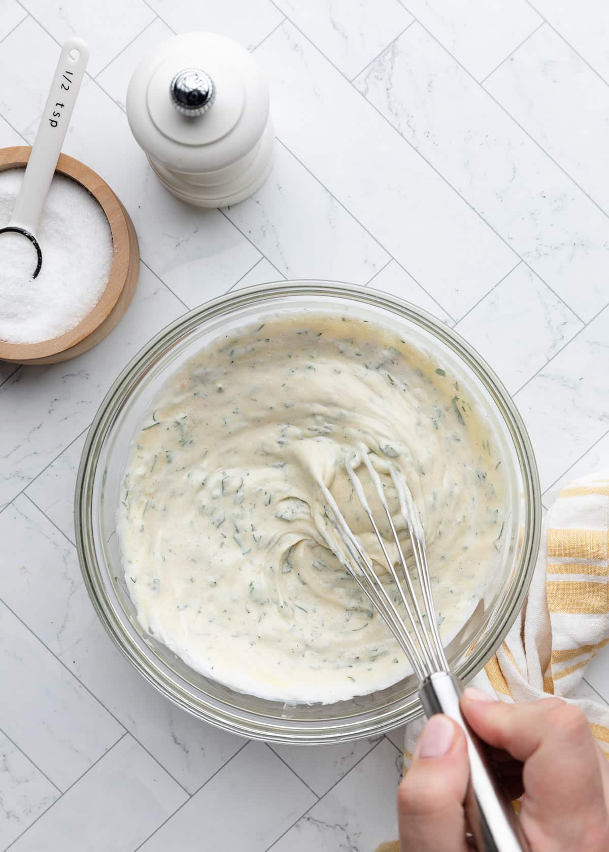 Overhead view of a creamy herb dressing being whisked together in a glass mixing bowl, with flecks of fresh herbs visible throughout and a striped kitchen towel beside the bowl on a white marble-patterned surface.