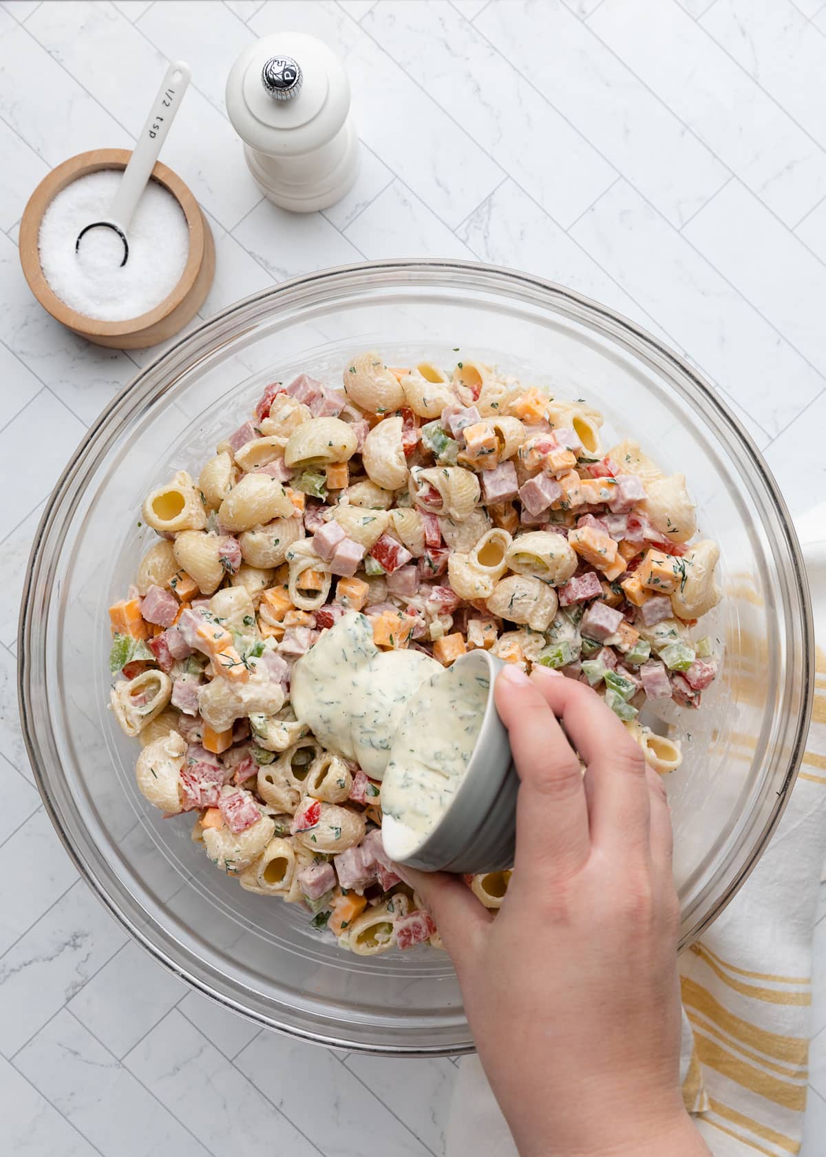 A hand pours extra creamy herb dressing from a small ramekin into a glass bowl of ham pasta salad made with shell pasta, diced ham, cheddar cheese, bell peppers, and fresh herbs, on a white marble-patterned surface with a salt cellar and kitchen towel nearby.