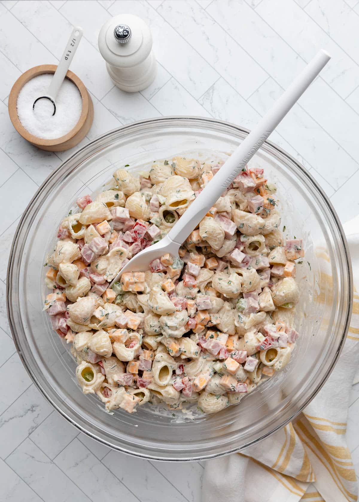 Overhead view of creamy ham pasta salad in a large glass mixing bowl with shell pasta, diced ham, cheddar cheese, red and green bell peppers, and herbs coated in a creamy dressing. A white serving spoon rests in the bowl beside a salt cellar, pepper mill, and striped kitchen towel.