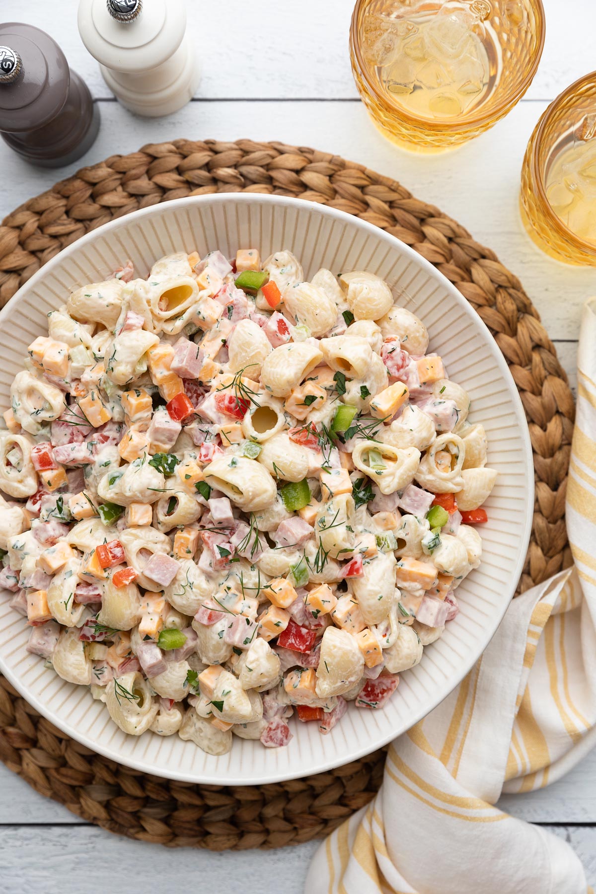 Overhead view of a large bowl of creamy ham pasta salad made with shell pasta, diced ham, cheddar cheese, red and green bell peppers, and fresh herbs, set on a woven placemat with iced drinks, salt and pepper mills, and a striped napkin.