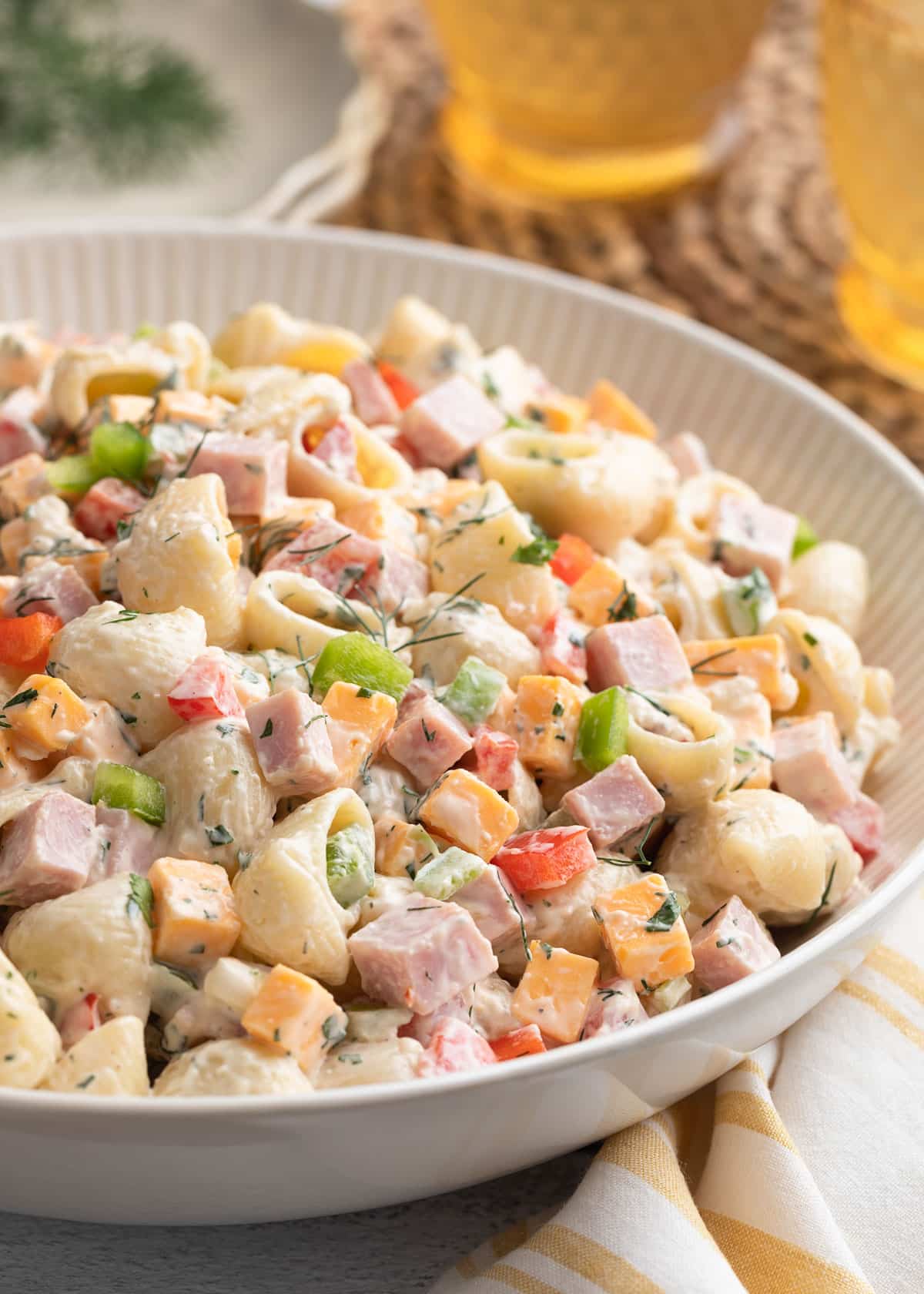 Close-up of creamy ham pasta salad with shell pasta, cubed ham, cheddar cheese, red and green bell peppers, and fresh dill in a white serving bowl, with iced drinks softly blurred in the background.