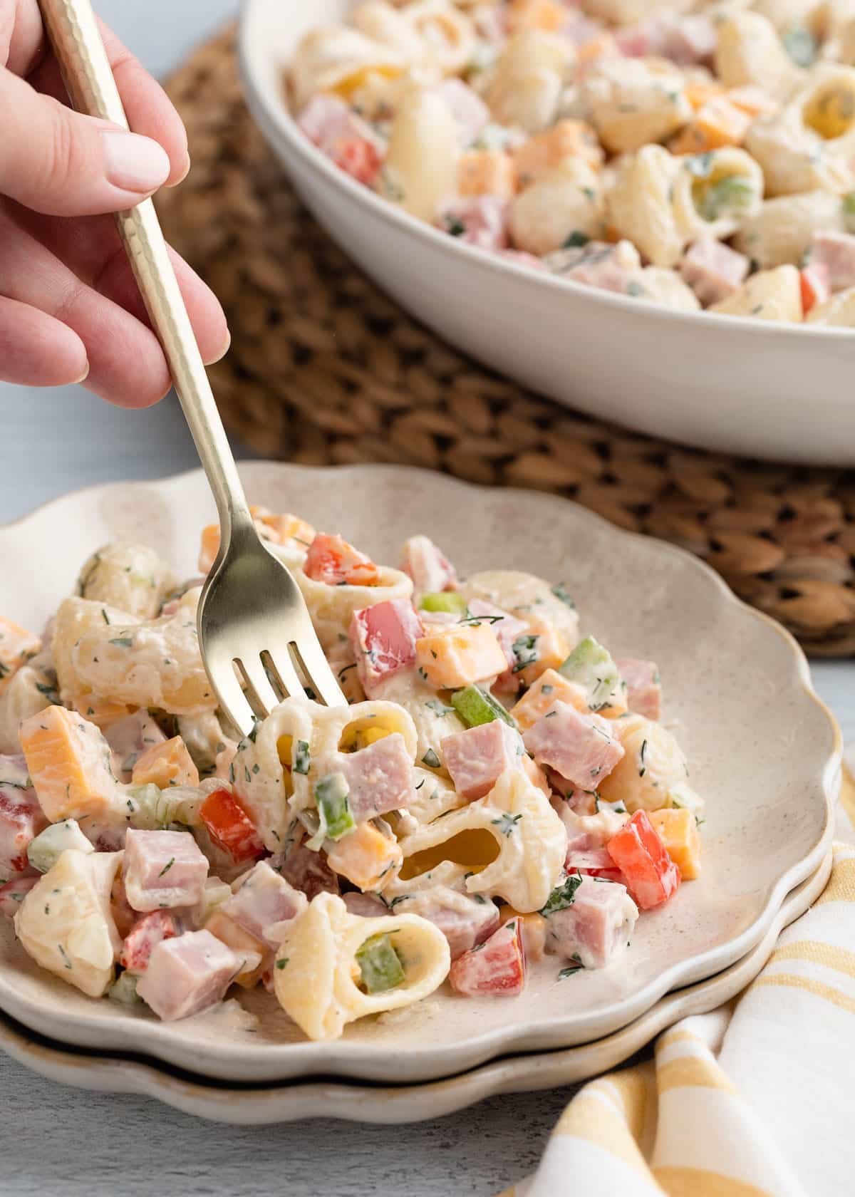 A fork lifts a serving of creamy ham pasta salad from a scalloped plate, showing shell pasta, diced ham, cheddar cheese, bell peppers, and herbs coated in a creamy dressing, with the serving bowl blurred behind.
