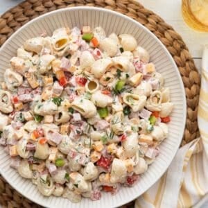Overhead view of a large bowl of creamy ham pasta salad made with shell pasta, diced ham, cheddar cheese, red and green bell peppers, and fresh herbs, set on a woven placemat with a striped napkin.