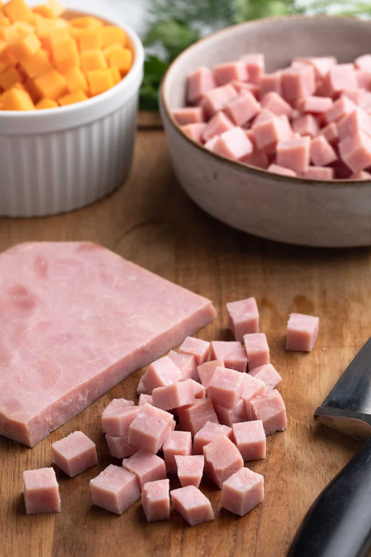 Close-up of diced ham on a wooden cutting board beside a thick slice of ham and a chef’s knife, with bowls of cubed cheddar cheese and additional diced ham blurred in the background.
