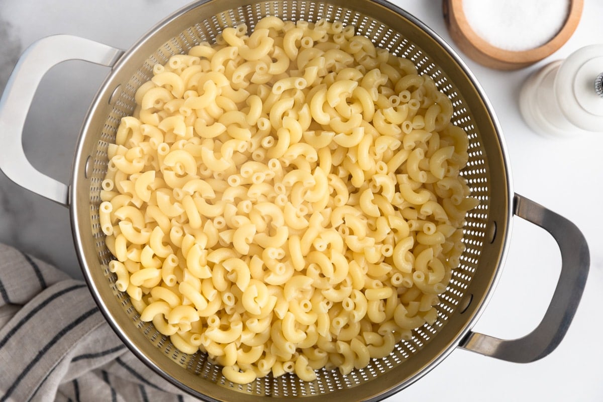 Cooked elbow macaroni draining in a stainless steel colander after boiling. The tender pasta is shown from above with a kitchen towel, salt cellar, and pepper mill nearby, illustrating a preparation step for homemade macaroni salad.