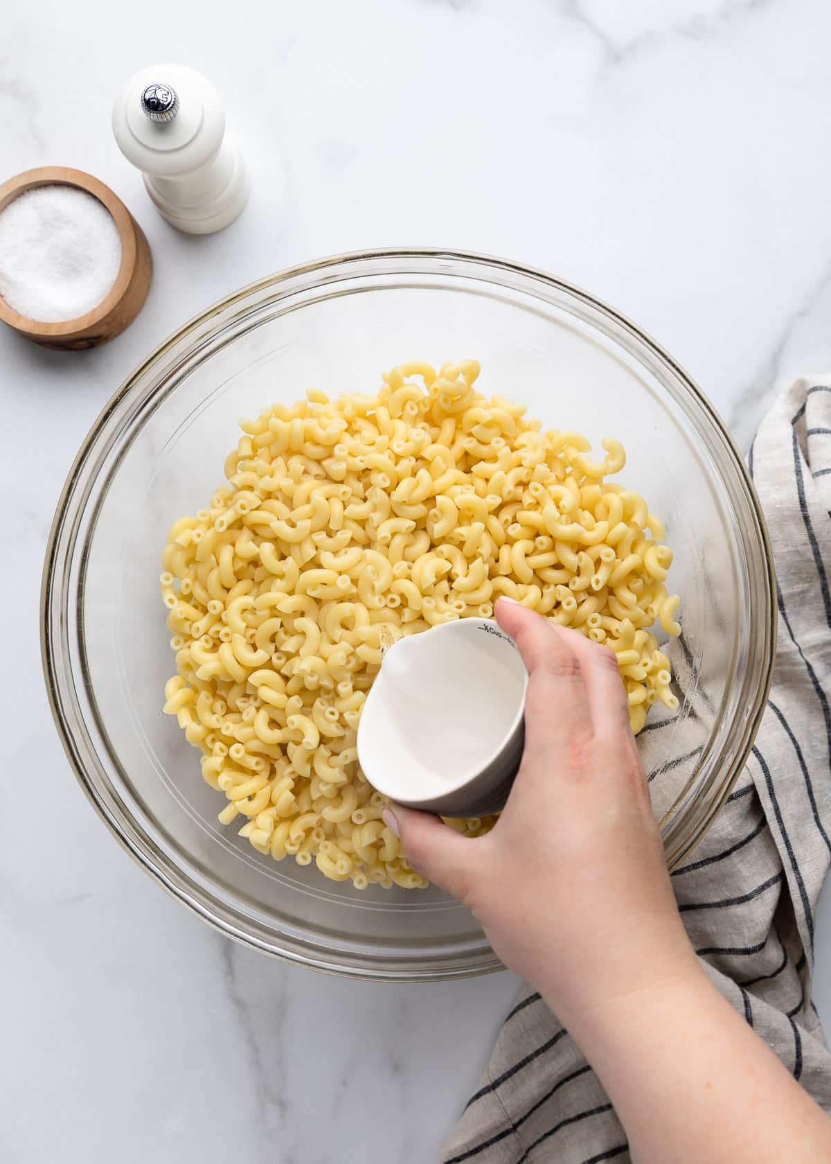 A hand pours distilled white vinegar over cooled elbow macaroni in a large glass bowl, beginning the dressing process for homemade macaroni salad. Salt, pepper, and a striped kitchen towel are arranged alongside the bowl.