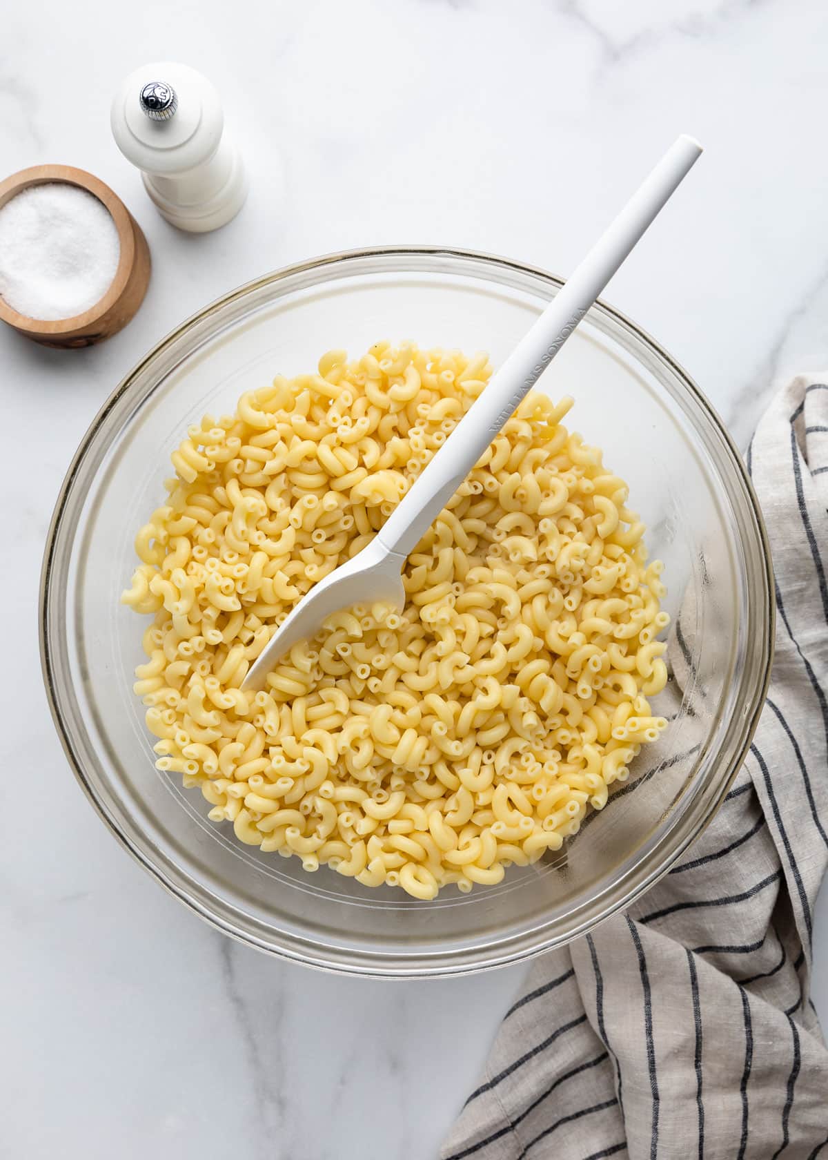 Overhead view of cooked elbow macaroni tossed with white vinegar in a large glass mixing bowl with a white rubber spatula. A salt cellar, pepper mill, and striped kitchen towel sit nearby.