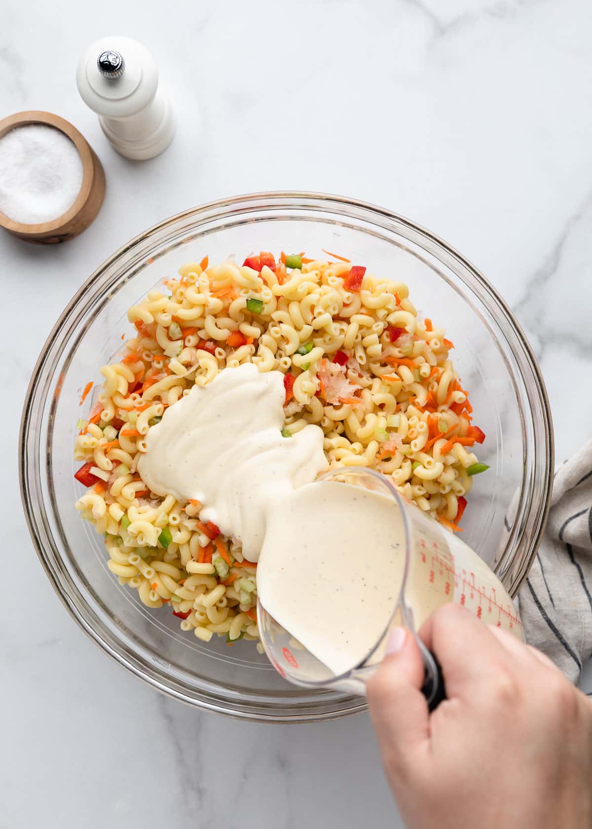 Creamy macaroni salad dressing being poured over cooked elbow macaroni mixed with carrots, celery, onion, and diced red and green bell peppers in a large glass bowl.