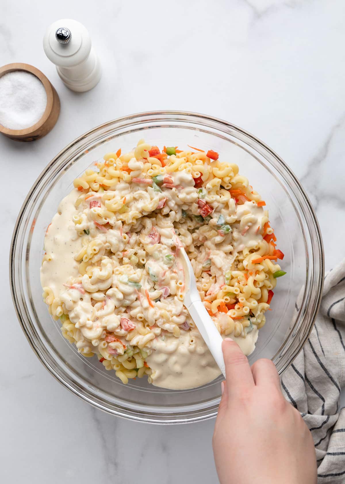 Cooked elbow macaroni coated in a creamy homemade dressing with grated carrots, diced celery, onion, and bell peppers, being stirred together in a large glass bowl to make classic macaroni salad.