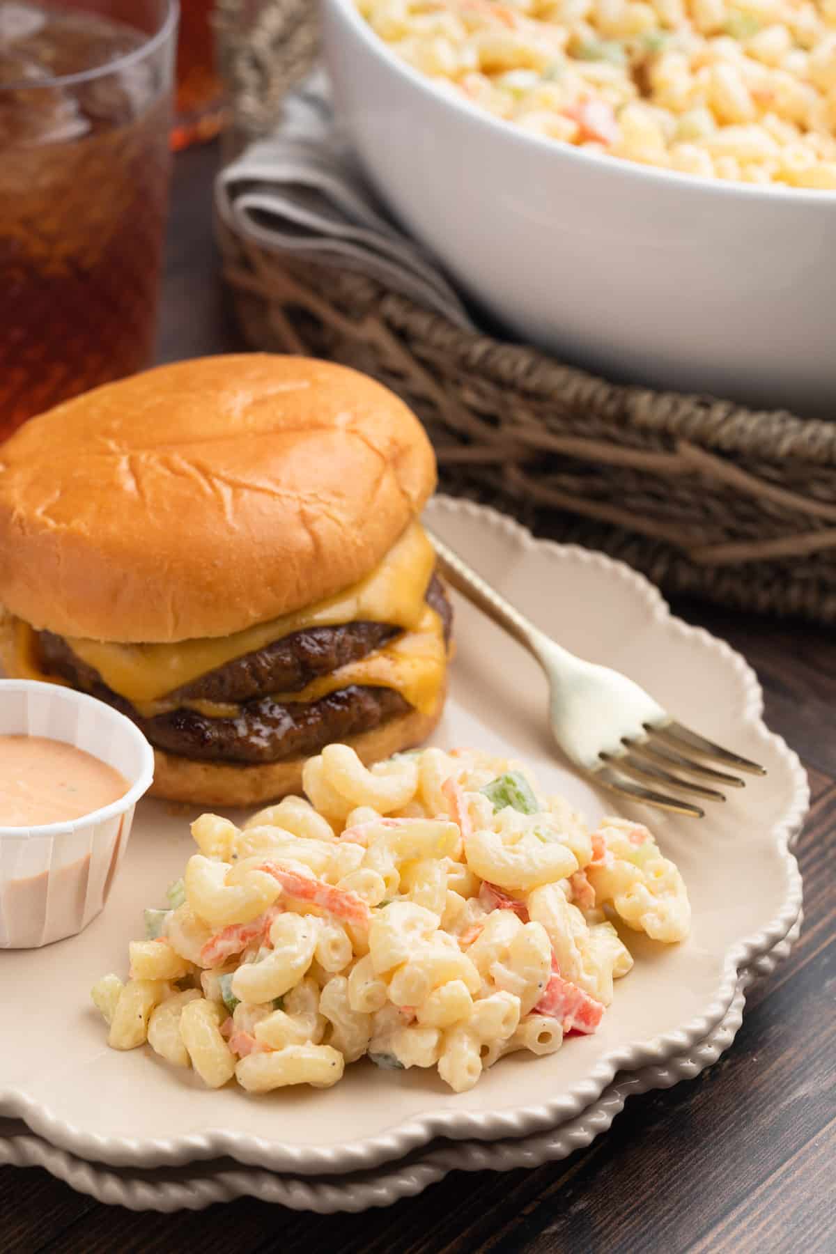 A cheeseburger topped with melted cheese is served on a plate alongside a scoop of creamy macaroni salad with diced vegetables. A small cup of pink dipping sauce sits beside the burger, while a large bowl of macaroni salad and iced drinks are visible in the background.