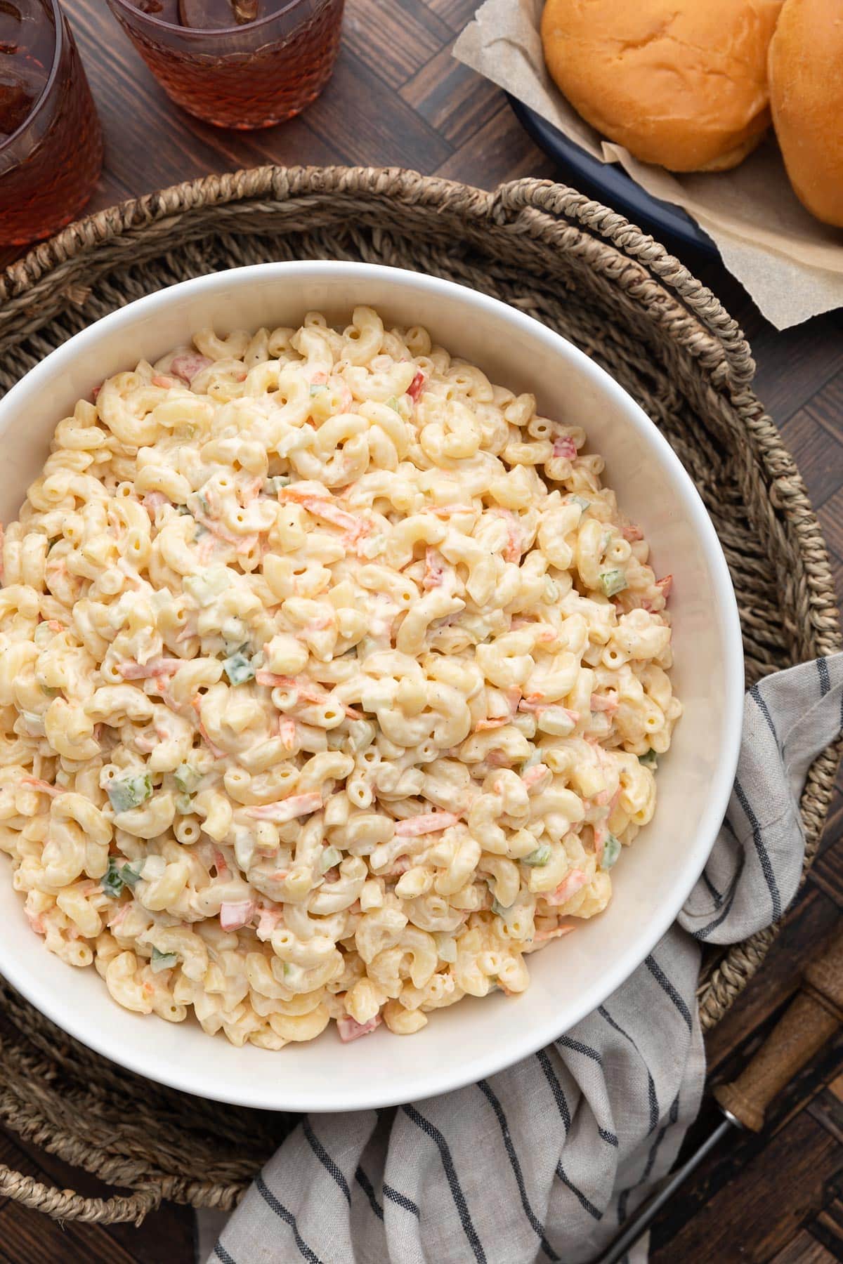 Overhead view of a large bowl of creamy macaroni salad filled with elbow pasta, shredded carrots, diced celery, and ham in a rich dressing. The bowl sits on a woven tray with burger buns, iced drinks, a striped kitchen towel, and grilling tools nearby, creating a casual cookout-style serving scene.