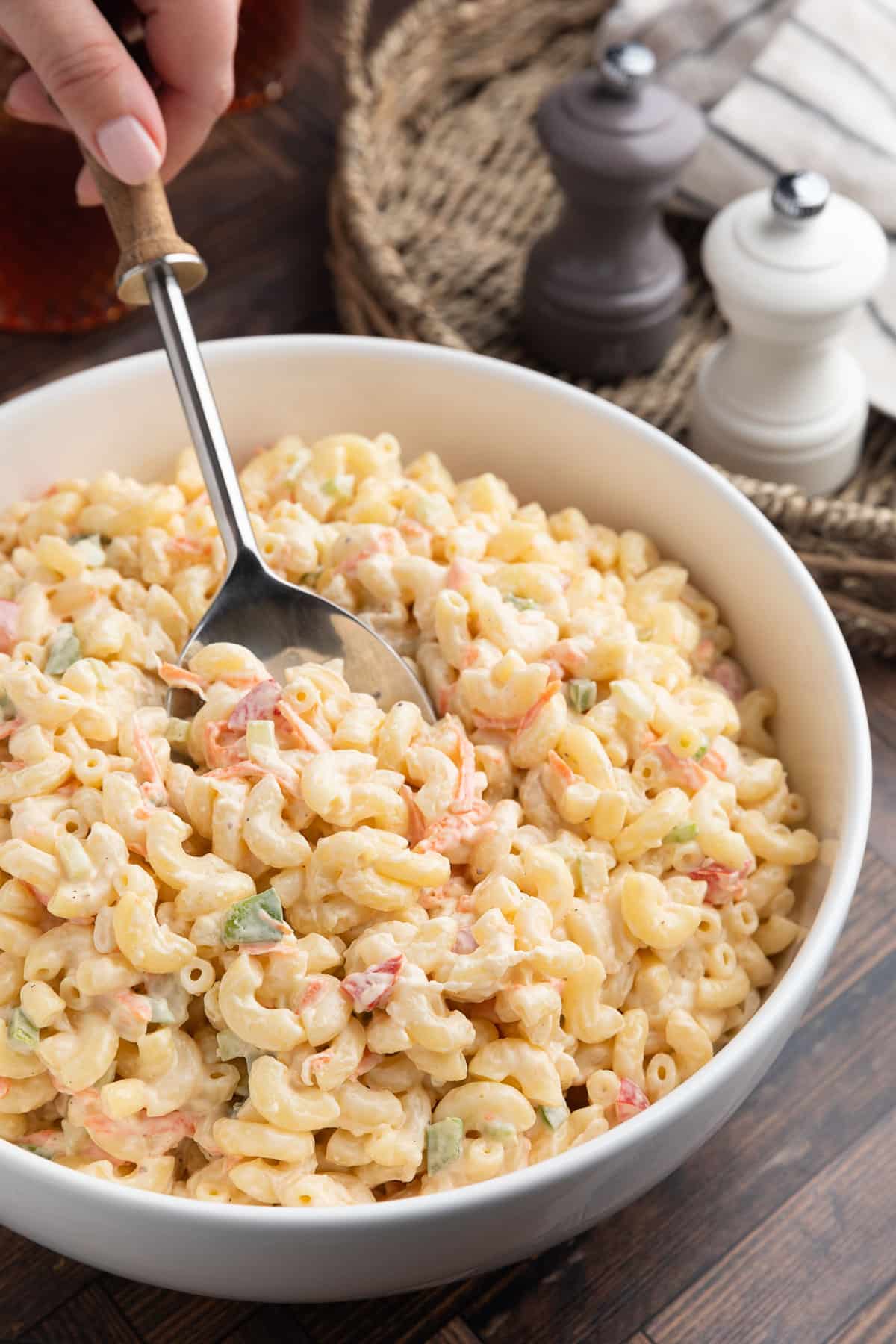 Creamy macaroni salad in a white serving bowl with a spoon scooping through the pasta. The salad is made with elbow macaroni, shredded carrots, diced celery, onion, and red and green bell peppers in a tangy mayonnaise dressing. Salt and pepper mills sit in the background on a dark wood tabletop.