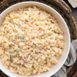 Close-up of creamy macaroni salad in a white serving bowl, featuring elbow macaroni coated in a mayonnaise-based dressing with shredded carrots, diced celery, onion, and bell peppers. The bowl sits on a woven tray with a striped kitchen towel beside it on a dark wood tabletop.